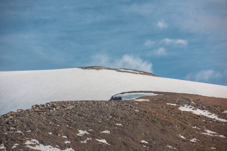 Dramatic landscape with huge rock mountain with snowy mountain dome in sunlight under clouds in blue sky. Awesome scenery with sunlit high snow mountain in dome shape in center at changeable weather.の写真素材