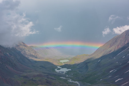 Bleak alpine landscape with vivid rainbow above mountain lake in valley. Gloomy scenery with bright rainbow above lake in mountain valley. Top view to colorful rainbow and low clouds in mountains.の写真素材