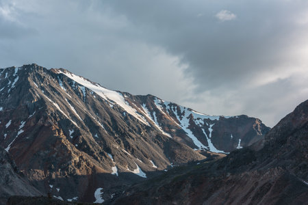 Dramatic overcast landscape with sunlit high mountain range with sharp rocks under gloomy sky. Dark atmospheric mountain scenery with large mountains in sunlight under cloudy sky at changeable weatherの写真素材