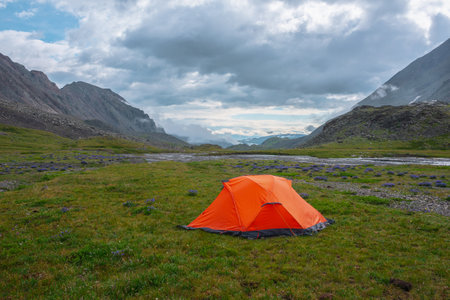 Scenic mountain landscape with tent in rain drops in sunlight under cloudy sky. Dramatic alpine scenery with vivid orange tent on grass among purple flowers in green mountain valley in rainy weather.の写真素材