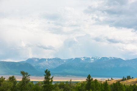 Dramatic view from forest to high mountain range in sunlight during rain in changeable weather. Colorful landscape with green forest and sunlit steppe against large mountains under cloudy sky in rain.の写真素材
