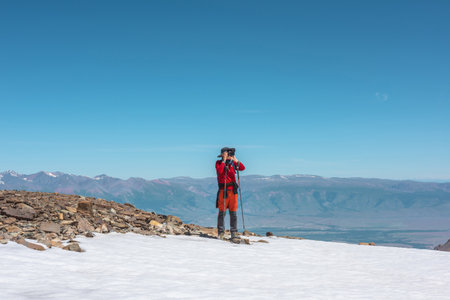 Tourist in red walks on snow mountain near abyss edge on high altitude under blue sky in sunny day. Man with camera on snowy mountain near precipice edge with view to large mountain range in away.の写真素材