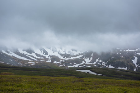 Dramatic alpine landscape with snowy mountains in gray low clouds. Bleak atmospheric scenery of tundra under lead gray sky. Gloomy minimalist view to mountain range among low rainy clouds in overcast.の写真素材