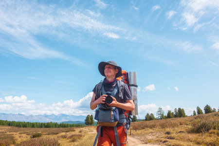 Alone traveler with large backpack walks along hiking trail on sunlit high mountain plateau under white clouds in blue sky. Backpacker with photo camera in autumn mountain trekking in good weather.の写真素材