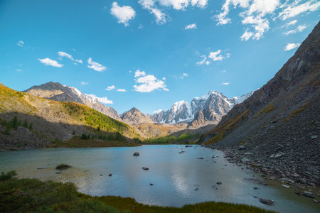 Pure azure alpine lake with view to forest hill and giant snow mountains in autumn sunny day. Glacial lake against huge snow covered mountain range in bright sun. Vivid autumn colors in high mountainsの写真素材