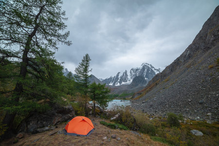 Dramatic landscape with alone orange tent on forest hill among rocks and autumn flora with view to large snowy mountain range under cloudy sky. Lonely tent and fading autumn colors in high mountains.の写真素材
