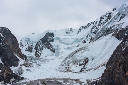 Dramatic landscape with large snow mountain top with ice cornice and glacier under gray cloudy sky. Big glacier with icefall on high altitude. Gloomy scenery in snowy mountains in overcast close-up.の写真素材