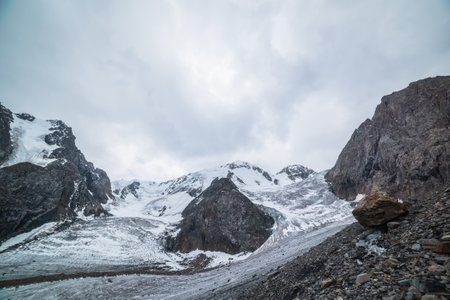 Dramatic landscape with two glacier icefalls on large snow mountain range with sharp rocks under gray cloudy sky. Long glacier with icefall in high altitude. Gloomy scenery in mountains in overcast.の写真素材