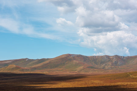 Motley autumn landscape with sunlit high mountain plateau and mountain range under dramatic cloudy sky. Vivid autumn colors in mountains. Sunlight and beautiful shadows of clouds in changeable weatherの写真素材