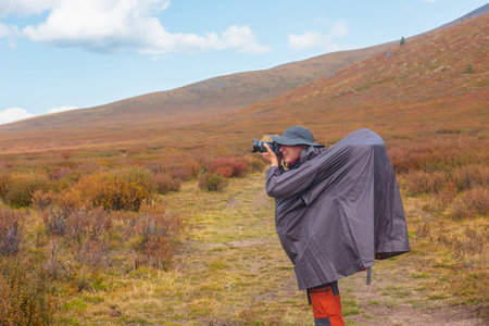 Alone traveler in raincoat with large backpack photographs nature among fading autumn vegetations on high mountain plateau. Backpacker with photo camera shoots mountain landscape in rainy weather.の写真素材