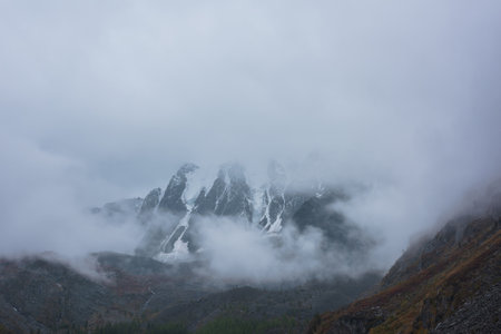 Dark atmospheric landscape with high snow mountains in dense fog in rainy weather. Large snowy mountain range in thick fog in dramatic overcast. Snow-covered rocky mountains in low clouds during rain.の写真素材