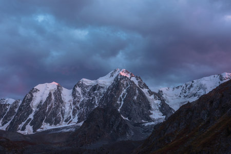 Awesome landscape with sunset pink reflection on huge snowy mountain top in violet dramatic sky. Hanging glacier and cornice on giant snow mountains in dusk. Snow-covered mountain range in twilight.の写真素材