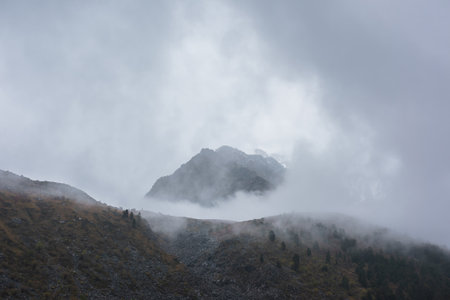 Dark atmospheric landscape with high mountain silhouettes in dense fog in rainy weather. Snowy rocky mountain top above hills in thick fog in dramatic overcast. Black rocks in low clouds during rain.の写真素材