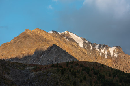 Awesome view to large mountain range and snowy peaked top among sharp rocks in golden sunlight under cloudy sky. Sunlit gold high mountain top with snow under dramatic gray sky in changeable weather.の写真素材