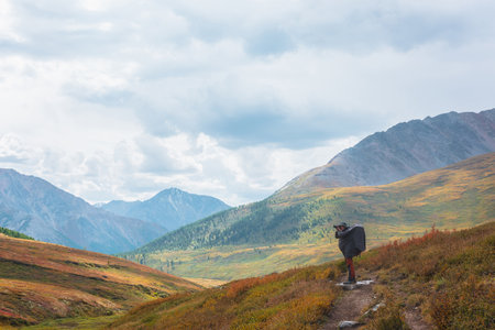 Alone traveler in raincoat with large backpack photographs nature among fading autumn vegetations on hiking trail through mountain pass. Backpacker with photo camera shoots autumn mountain landscape.の写真素材