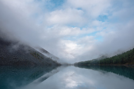Tranquil meditative misty scenery of glacial lake with reflection of pointy fir tops and clouds at early morning. Graphic EQ of spruce silhouettes on calm alpine lake horizon. Mountain lake in fog.の写真素材