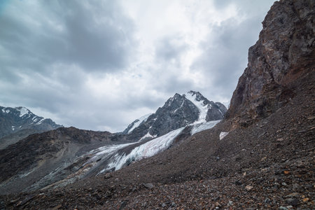 Dramatic landscape with hanging glacier against large snow mountain with sharp rocky pinnacle under gray cloudy sky. Glacier with icy cornice in high altitude. Gloomy scenery in mountains in overcast.の写真素材