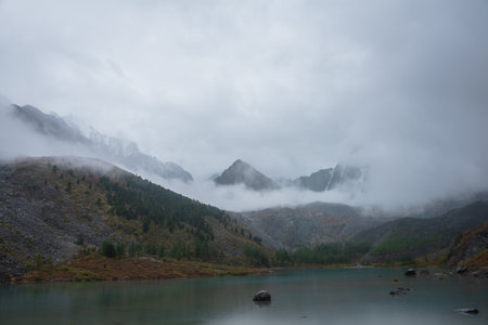 Dark atmospheric landscape with alpine lake and high mountain silhouettes in dense fog. Snowy rocky mountain top above hills in thick fog in dramatic overcast. Black rocks in low clouds during rain.の写真素材