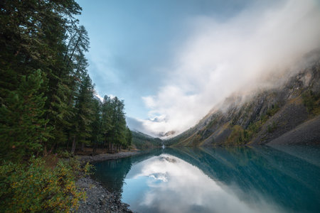 Scenic view to big pure mountain lake in autumn valley against snow mountain top in low clouds....