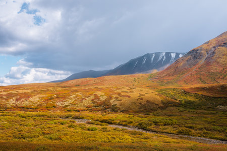 Motley autumn landscape with sunlit hills and mountain range silhouette under dramatic cloudy sky. Vivid autumn colors in mountains. Sunlight on multicolor hills and rainy clouds in changeable weatherの写真素材