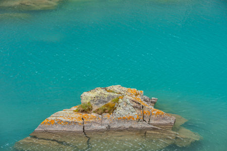 Stone with grass and lichens in azure transparent water of mountain lake. Fragment of meditative lake with lichen rock. Scenic nature background of relaxing shiny ripples on calm water of alpine lake.の写真素材