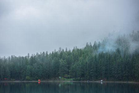 Tranquil atmospheric scenery with tents near mountain lake among coniferous trees silhouettes in dense fog. Pure alpine lake in mystery thick fog. Glacial lake and forest edge in misty early morning.の写真素材