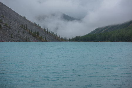 Dramatic meditate landscape with ripples on big azure mountain lake against pointy firs silhouettes in low clouds. Tranquil view to cyan alpine lake and peaked tops of spruces in low cloudy gray sky.の写真素材