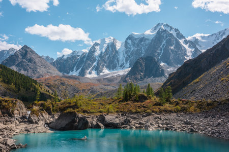 Pure turquoise alpine lake with view to forest hill and giant snow mountains in autumn sunny day. Glacial lake against huge snow covered mountain range in bright sun. Vivid colors in high mountains.の写真素材