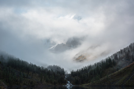 Tranquil scenery with snow castle in clouds and alpine lake. Mountain creek flows from forest hills into glacial lake. Snowy mountains in fog clearance. Small river and coniferous trees in low clouds.の写真素材