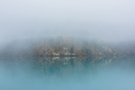 Simple meditative landscape with turquoise lake in thick fog. Tranquil view through clearance in dense fog to steep slope in fading autumn colors and mirror mountain lake. Reflective azure alpine lakeの写真素材