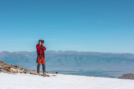 Tourist in red walks on snow mountain near abyss edge on high altitude under blue sky in sunny day. Man with camera on snowy mountain near precipice edge with view to large mountain range in away.の写真素材