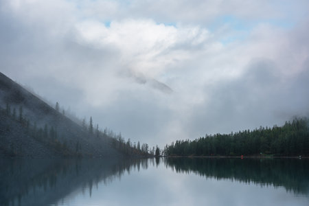 Tranquil meditative misty scenery of glacial lake with reflection of pointy fir tops and clouds at early morning. Graphic EQ of spruce silhouettes on calm alpine lake horizon. Mountain lake in fog.の写真素材