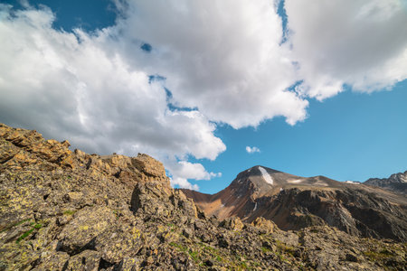 Scenic landscape with sunlit stone hill and mountain top with snow under cloudy sky. Colorful scenery with sharp rocks and mountain range in sunlight under clouds in blue sky at changeable weather.の写真素材