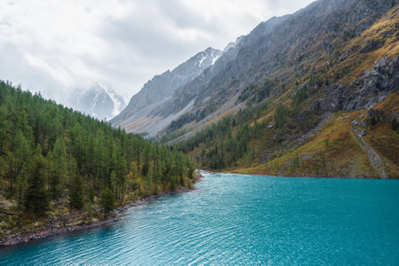 Shiny ripples on turquoise alpine lake against snowy mountain range during rain in changeable weather. Big azure mountain lake in forest valley in vivid autumn colors and snow mountains in overcast.の写真素材