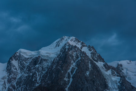Atmospheric nightly landscape with huge snowy mountain top in dramatic sky. Hanging glacier and cornice on beautiful giant snow mountains in night. High snow-covered mountain range in dusk dim light.の写真素材