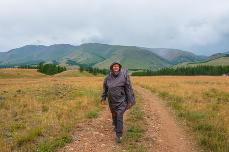 Satisfied tourist returns from mountains in overcast. Happy man in raincoat walks through hills and forest in bad weather. Traveler goes towards adventure. Hiker and mountain range under cloudy sky.の写真素材