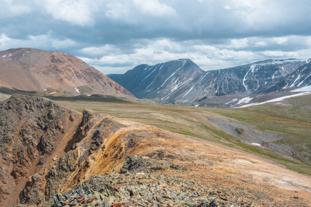 Dramatic alpine landscape with abyss and high mountain range in sunlight under cloudy sky. Colorful scenery with sunlit sharp rocks near precipice edge and large mountain ridge at changeable weather.の写真素材