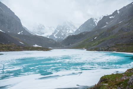 Atmospheric mountain landscape with frozen alpine lake during snowfall. Awesome cold scenery with snowfall in high mountain valley with icy mountain lake on background of snow mountains in low clouds.の写真素材