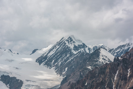 Atmospheric landscape with sharp rocks and high snowy mountain top in rainy low clouds at overcast. Dramatic gloomy scenery with large snow mountains and glacier in gray cloudy sky at rainy weather.の写真素材