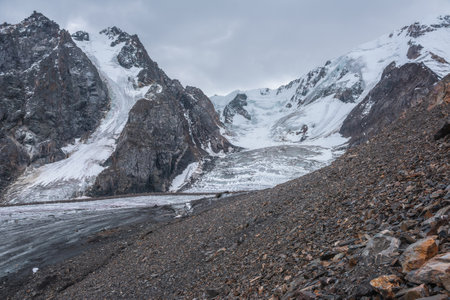 Dramatic landscape with glacier icefall on large snow mountain range with sharp rocky pinnacle under gray cloudy sky. Big glacier close-up in high altitude. Gloomy scenery in mountains in overcast.の写真素材