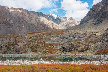 Turquoise mountain lake among autumn multicolor flora on rocks against high rocky mountain top in sunlight under clouds in blue sky. Sunlit alpine lake and large mountain range. Vivid autumn colors.の写真素材