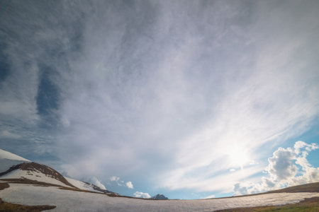 Dramatic landscape with sunlit snowy mountain ridge against pointed peak with snow in sunlight in cloudy sky in changeable weather. Colorful sunny scenery with glacier and snow mountain peaked top.の写真素材
