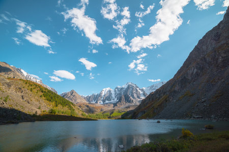 Pure azure alpine lake with view to forest hill and giant snow mountains in autumn sunny day. Glacial lake against huge snow covered mountain range in bright sun. Vivid autumn colors in high mountainsの写真素材