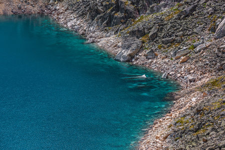 Top view to azure mountain lake with shiny cyan water edge among sunlit rocks. Beautiful alpine lake with stony bottom in transparent water in bright sun. Nature background of blue lake in sunlight.の写真素材