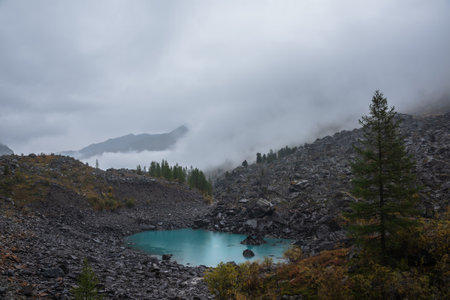 Dark atmospheric landscape with turquoise alpine lake against stone hill with forest and silhouette of mountain range in dense fog. Small mountain lake and black rocks in thick low clouds during rain.の写真素材