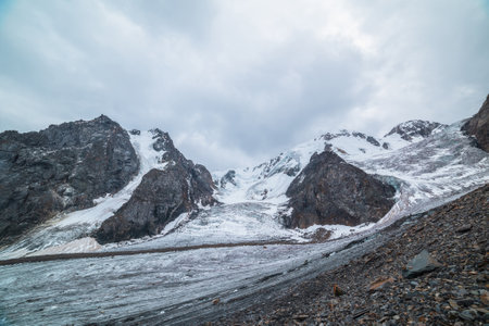 Dramatic landscape with two glacier icefalls on large snow mountain range with sharp rocks under gray cloudy sky. Long glacier with icefall in high altitude. Gloomy scenery in mountains in overcast.の写真素材