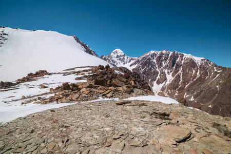 Scenic mountain landscape with sunlit stone outliers and high snow mountain with peaked top under blue sky in sunny day. Awesome alpine scenery with sharp rocks and high snowy pinnacle in sunlight.の写真素材