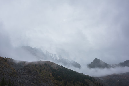 Dark atmospheric landscape with high mountain silhouettes in dense fog in rainy weather. Snowy rocky mountain top above hills in thick fog in dramatic overcast. Black rocks in low clouds during rain.の写真素材