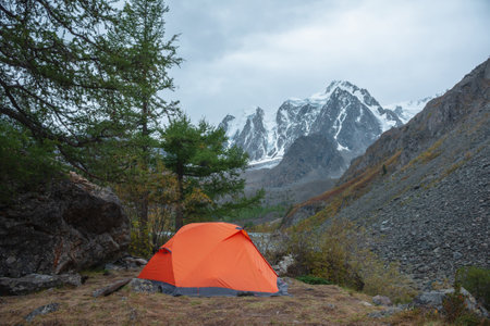 Dramatic landscape with alone orange tent on forest hill among rocks and autumn flora with view to large snowy mountain range under cloudy sky. Lonely tent and fading autumn colors in high mountains.の写真素材