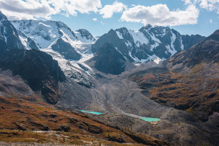 Motley autumn landscape with two small lakes in high valley and large snow mountain range with glacier in sunny day. Vivid autumn colors in mountains. Glacial lakes and mountain wall in bright sun.の写真素材
